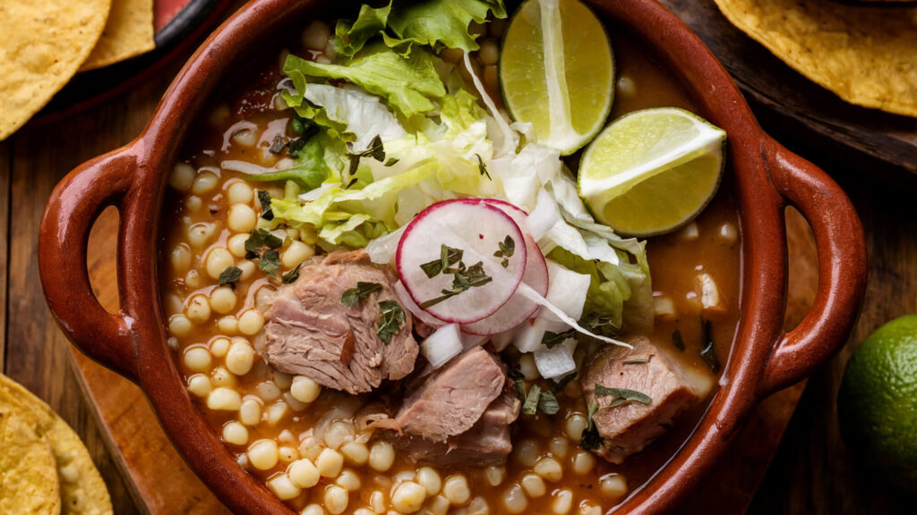 A bowl of Pozole Rojo filled with pork, hominy, and red chile broth, topped with cabbage, radishes, and lime.